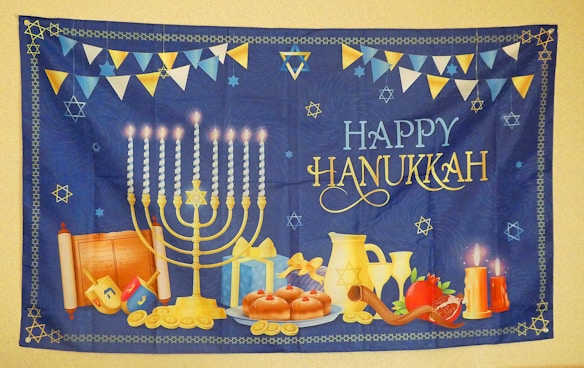 A festive Hanukkah tapestry featuring a lit menorah with nine candles, surrounded by traditional items like dreidels, donuts, a gift box, coins, and a shofar. A pair of lit candles, a pomegranate, and a Torah scroll add to the scene. 'Happy Hanukkah' is prominently displayed in decorative lettering. The background is deep blue with a border and scattered Star of David motifs, accompanied by bunting flags in blue, white, and yellow.