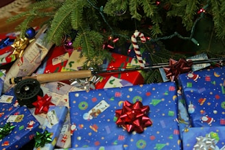 A collection of wrapped presents with colorful bows sits beneath a Christmas tree. The wrapping paper features festive designs such as snowmen, Santa Claus, and ornaments. A fishing rod with a reel is also present among the gifts, indicating it might be a Christmas gift. The tree branches are adorned with ornaments and lights.