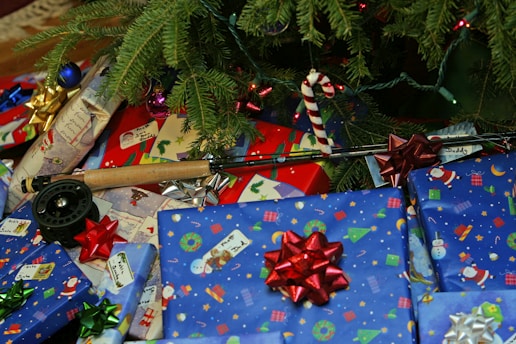 A collection of wrapped presents with colorful bows sits beneath a Christmas tree. The wrapping paper features festive designs such as snowmen, Santa Claus, and ornaments. A fishing rod with a reel is also present among the gifts, indicating it might be a Christmas gift. The tree branches are adorned with ornaments and lights.