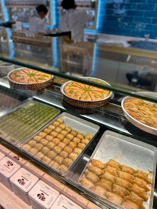 An array of various types of baklava is displayed in a glass case, including different shapes and sizes, with some garnished with pistachios. Behind the counter, two people wearing white uniforms are preparing or managing food in a brightly lit environment with a blue-tiled wall.