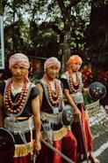 Three men wearing traditional attire with headscarves, necklaces made of large beads, and woven belts with metallic details are holding round shields and spears. They stand outdoors, surrounded by lush greenery and plants.