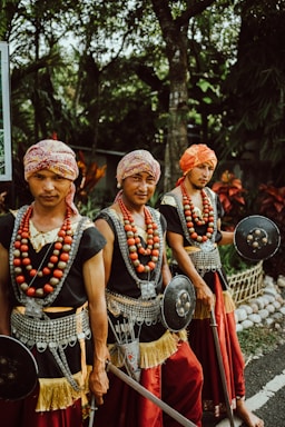 Three men wearing traditional attire with headscarves, necklaces made of large beads, and woven belts with metallic details are holding round shields and spears. They stand outdoors, surrounded by lush greenery and plants.