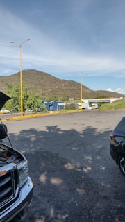 A parking area with several vehicles, including a truck and a few cars, is situated in front of a mountainous landscape. The sky is mostly clear with a few clouds, and there are yellow lamp posts lining the road. There is some shadow on the ground indicating sunlight filtering through trees nearby.
