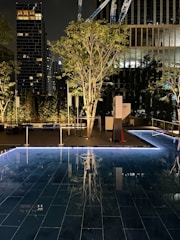 A night-time scene featuring a rooftop pool with calm, reflective water. The poolside area is surrounded by trees and illuminated by soft, ambient lighting. In the background, tall buildings with lit windows are visible, along with construction cranes indicating ongoing development.