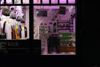 A store interior with neatly arranged clothing displayed on hangers, including tie-dye and graphic tees. Skateboard decks and various merchandise on shelves and in glass display cases. A checkerboard floor and subdued lighting with purple hues create a cozy atmosphere.