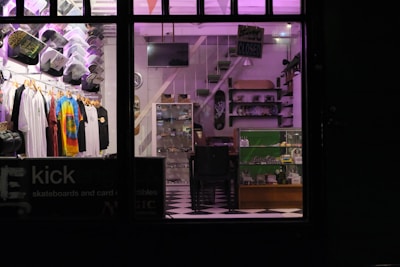 A store interior with neatly arranged clothing displayed on hangers, including tie-dye and graphic tees. Skateboard decks and various merchandise on shelves and in glass display cases. A checkerboard floor and subdued lighting with purple hues create a cozy atmosphere.