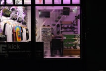 A store interior with neatly arranged clothing displayed on hangers, including tie-dye and graphic tees. Skateboard decks and various merchandise on shelves and in glass display cases. A checkerboard floor and subdued lighting with purple hues create a cozy atmosphere.