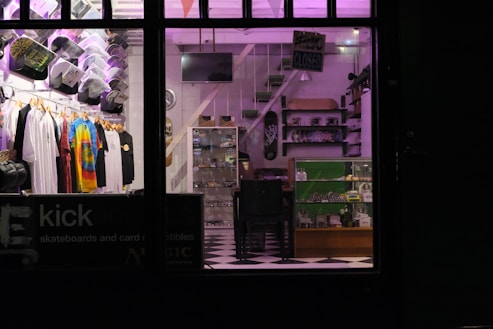 A store interior with neatly arranged clothing displayed on hangers, including tie-dye and graphic tees. Skateboard decks and various merchandise on shelves and in glass display cases. A checkerboard floor and subdued lighting with purple hues create a cozy atmosphere.