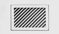 A rectangular, white air vent is mounted on a white wall. The vent has diagonal slats that create a dynamic pattern of shadows and light.
