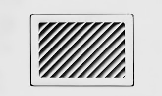 A rectangular, white air vent is mounted on a white wall. The vent has diagonal slats that create a dynamic pattern of shadows and light.