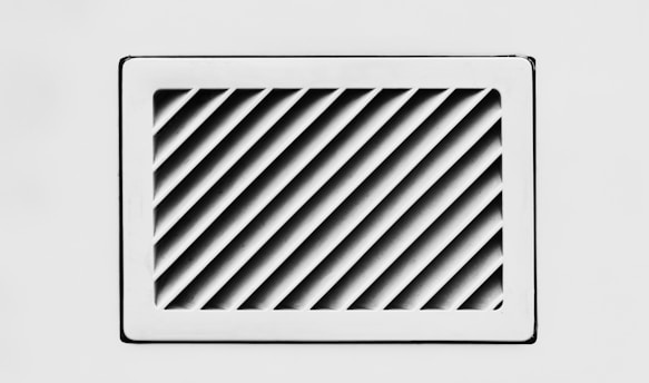 A rectangular, white air vent is mounted on a white wall. The vent has diagonal slats that create a dynamic pattern of shadows and light.