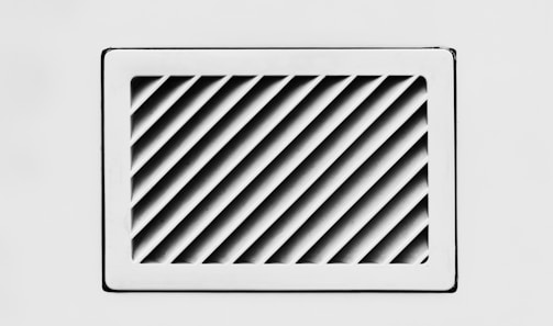 A rectangular, white air vent is mounted on a white wall. The vent has diagonal slats that create a dynamic pattern of shadows and light.