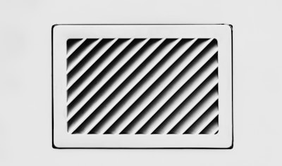 A rectangular, white air vent is mounted on a white wall. The vent has diagonal slats that create a dynamic pattern of shadows and light.