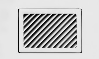 A rectangular, white air vent is mounted on a white wall. The vent has diagonal slats that create a dynamic pattern of shadows and light.
