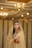A bride adorned in a traditional gold outfit with intricate jewelry, including a headpiece and earrings, standing in an elegantly decorated indoor setting. Her hands and arms are decorated with henna, and she wears a delicate veil that partially covers her face. The background features warm lighting with chandeliers, enhancing the opulent atmosphere.