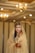 A bride adorned in a traditional gold outfit with intricate jewelry, including a headpiece and earrings, standing in an elegantly decorated indoor setting. Her hands and arms are decorated with henna, and she wears a delicate veil that partially covers her face. The background features warm lighting with chandeliers, enhancing the opulent atmosphere.