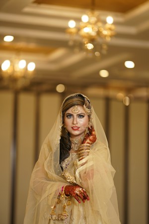 A bride adorned in a traditional gold outfit with intricate jewelry, including a headpiece and earrings, standing in an elegantly decorated indoor setting. Her hands and arms are decorated with henna, and she wears a delicate veil that partially covers her face. The background features warm lighting with chandeliers, enhancing the opulent atmosphere.