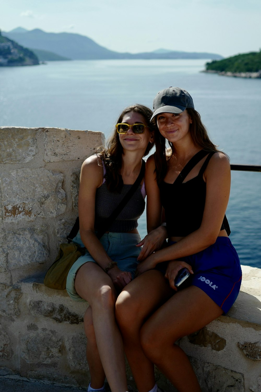 two women sitting on a stone wall next to a body of water
