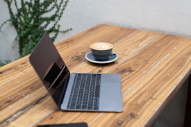 A wooden table with a laptop and a cup of cappuccino placed on it. The cappuccino has a decorative latte art on top, and the table's surface features visible wood grain and natural imperfections. Some green plants are visible in the background.