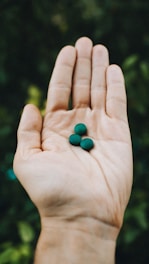 A hand is open with three small, round green pills or beads resting on the palm against a blurred, leafy background.