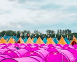 A campsite filled with numerous vibrant tents, predominantly purple in the foreground, with yellow and blue teepee-style tents in the background. The site is decorated with colorful flags perched atop some of the tents and surrounded by lush green trees under an overcast sky.