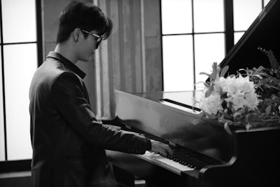 A person wearing sunglasses and a suit is playing a grand piano in a well-lit room with large windows. There are white flowers and green foliage resting on the piano.