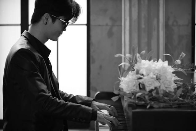 A person wearing sunglasses and a leather jacket is playing the piano in an elegant room with large windows. A bouquet of white flowers rests on top of the piano.