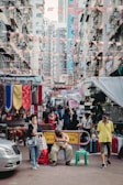 A bustling urban street market set between tall, closely-packed apartment buildings. Banners and flags are strung overhead, adding a festive touch. The street is lined with colorful textiles and stalls selling various goods like bags. A mix of people walk lazily around, one person sitting with a hunched posture, another standing while on the phone, and a third person wearing a hat walking by.