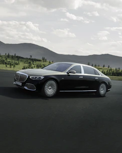A sleek black luxury sedan parked on a winding mountain road with pine trees and misty hills in the background.