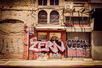 An old, weathered building facade is covered with graffiti, including the words 'ZERM' in bold, red letters. The building features a mix of brick and stone textures, with barred windows and rusted metal shutters. An air conditioning unit is mounted on the wall along with a number 21 sign.