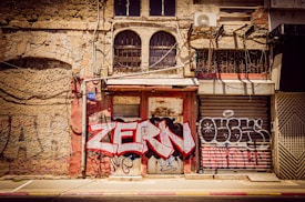 An old, weathered building facade is covered with graffiti, including the words 'ZERM' in bold, red letters. The building features a mix of brick and stone textures, with barred windows and rusted metal shutters. An air conditioning unit is mounted on the wall along with a number 21 sign.