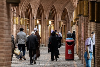 A bustling indoor market with arched brick ceilings and ornate lamps. People are walking, dressed in diverse attire, including traditional garments. A red waste bin and cobblestone flooring are visible.