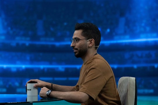 A person wearing glasses and a casual brown shirt is seated at a table with a microphone earpiece. There is a white cup on the table and a vibrant blue backdrop that suggests the setting of a sports stadium or studio.