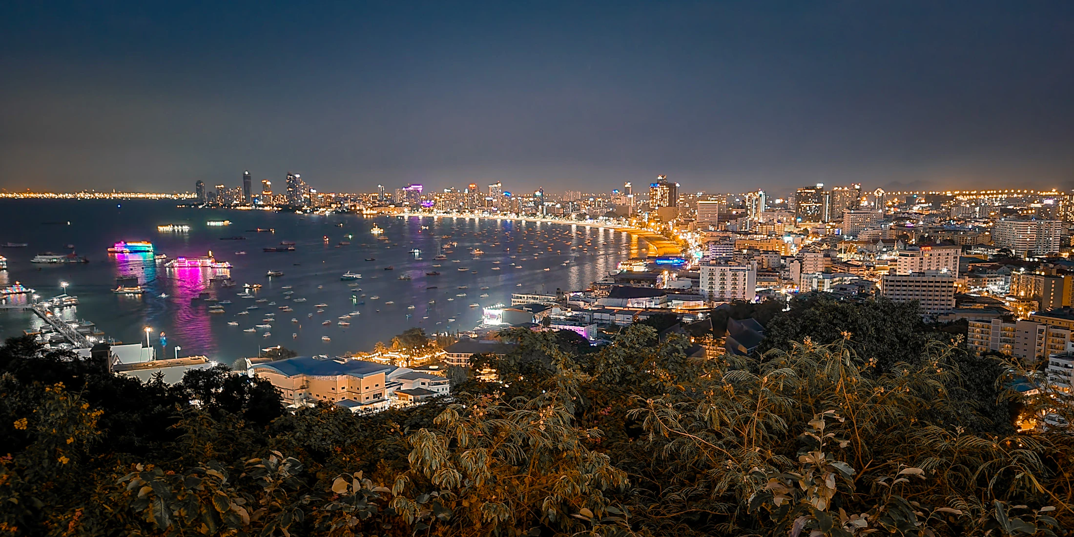 Pattaya bay at night seen from the hills — lit skyline, boats on dark water, the strip glowing