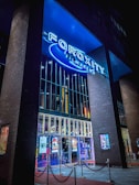A modern cinema entrance illuminated in blue neon lights. Large glass doors reflect the vibrant interior lights, while colorful movie posters adorn the exterior walls. The structure is framed by tall brick pillars, and red velvet ropes guide visitors inside.
