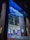 A modern cinema entrance illuminated in blue neon lights. Large glass doors reflect the vibrant interior lights, while colorful movie posters adorn the exterior walls. The structure is framed by tall brick pillars, and red velvet ropes guide visitors inside.