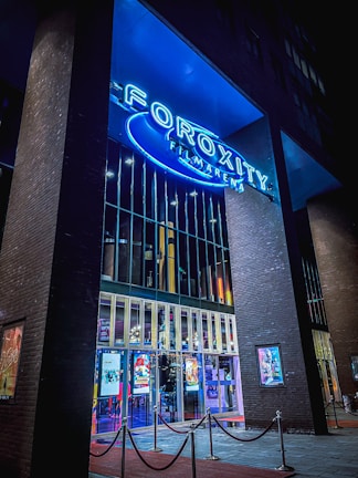 A modern cinema entrance illuminated in blue neon lights. Large glass doors reflect the vibrant interior lights, while colorful movie posters adorn the exterior walls. The structure is framed by tall brick pillars, and red velvet ropes guide visitors inside.