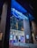 A modern cinema entrance illuminated in blue neon lights. Large glass doors reflect the vibrant interior lights, while colorful movie posters adorn the exterior walls. The structure is framed by tall brick pillars, and red velvet ropes guide visitors inside.