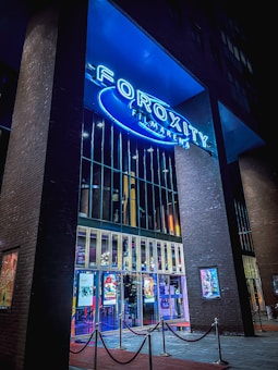 A modern cinema entrance illuminated in blue neon lights. Large glass doors reflect the vibrant interior lights, while colorful movie posters adorn the exterior walls. The structure is framed by tall brick pillars, and red velvet ropes guide visitors inside.