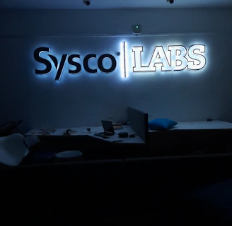 A dimly lit room features a prominent illuminated sign displaying the words 'Sysco LABS.' Below the sign are office desks with various electronic devices, including laptops and cables. The lighting creates a cool and professional atmosphere.