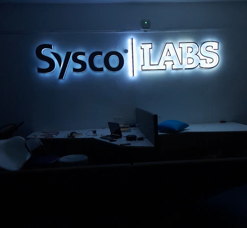 A dimly lit room features a prominent illuminated sign displaying the words 'Sysco LABS.' Below the sign are office desks with various electronic devices, including laptops and cables. The lighting creates a cool and professional atmosphere.