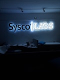 A dimly lit room features a prominent illuminated sign displaying the words 'Sysco LABS.' Below the sign are office desks with various electronic devices, including laptops and cables. The lighting creates a cool and professional atmosphere.