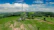 A telecommunications tower is situated in a vast, green rural landscape. The surrounding area consists of rolling hills dotted with trees and patches of open grassland. In the background, there's a clear blue sky with some clouds scattered across it, and a dirt road winds through the fields leading towards the tower.