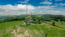 A telecommunications tower is situated in a vast, green rural landscape. The surrounding area consists of rolling hills dotted with trees and patches of open grassland. In the background, there's a clear blue sky with some clouds scattered across it, and a dirt road winds through the fields leading towards the tower.
