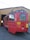 A red tram with yellow accents is parked partially inside a wooden garage. The tram has several rectangular windows and a pantograph on top. Bright yellow signage with black text is visible on its side, and the number 210 is displayed prominently at the rear. The surrounding area is a paved surface leading into the garage.