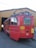 A red tram with yellow accents is parked partially inside a wooden garage. The tram has several rectangular windows and a pantograph on top. Bright yellow signage with black text is visible on its side, and the number 210 is displayed prominently at the rear. The surrounding area is a paved surface leading into the garage.
