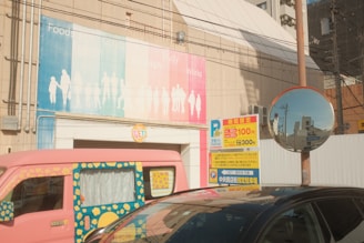 A colorful van with a unique design featuring yellow flowers and blue accents is parked near a street sign displaying parking information with prices in Japanese yen. A convex mirror is mounted on a pole, reflecting part of the surrounding environment, including buildings and the sky. In the background, a large billboard on a building wall shows silhouettes of people with text sections divided into colors like blue, green, and pink. Various industrial and urban architectural elements are visible, including power lines and building structures.