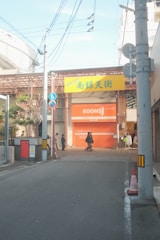A street leads to an entrance with a yellow sign containing Japanese characters. Below, a closed orange shutter with the word 'ROOMS' is visible. Several people walk along the sidewalk, and power lines cross above. There are traffic cones and street signs also present.