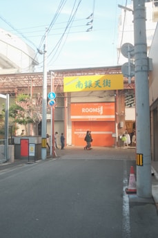 A street leads to an entrance with a yellow sign containing Japanese characters. Below, a closed orange shutter with the word 'ROOMS' is visible. Several people walk along the sidewalk, and power lines cross above. There are traffic cones and street signs also present.