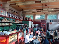 A bustling cafe interior with people sitting at tables, engaged in conversations and enjoying their meals. The decor features green tiles, wooden elements, and traditional designs on the walls and ceiling, creating a warm and inviting atmosphere. A display case filled with bakery items and a counter where staff are serving customers are visible on the left side.